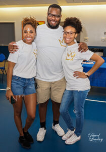 Three smiling adults wearing a white t-shirt with a gold crown on it.