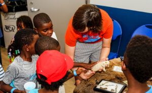 Children gathered around an adult who is holding a reptile for them to see and pet.