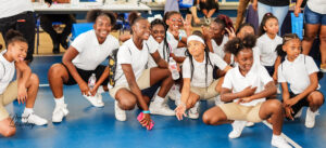 A group of children all dressed in white shirts and shorts, sitting on a gymnasium floor, posing for the camera.